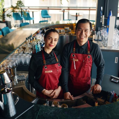 “Two bartenders in aprons smiling behind a bar in a modern restaurant”