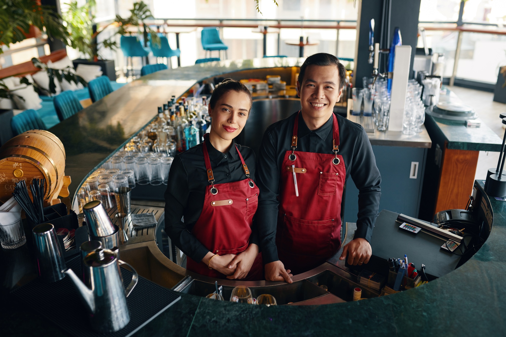 “Two bartenders in aprons smiling behind a bar in a modern restaurant”
